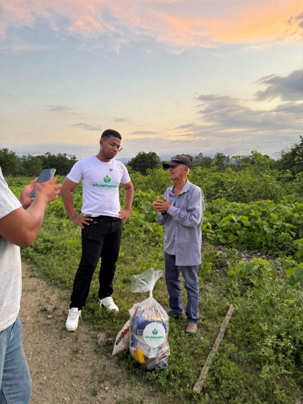 A Wholehuman team member talking with an older man beside a bag of food in a field at sunset