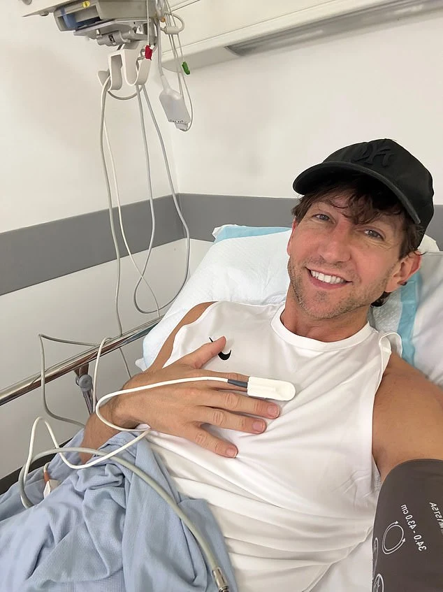 Ross smiling from a hospital bed, wearing a white shirt and black cap